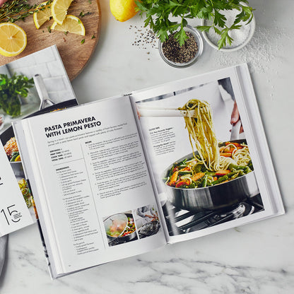 Cookbook open to Pasta Primavera recipe on marble counter, featuring a photo of noodles and vegetables in a pan.