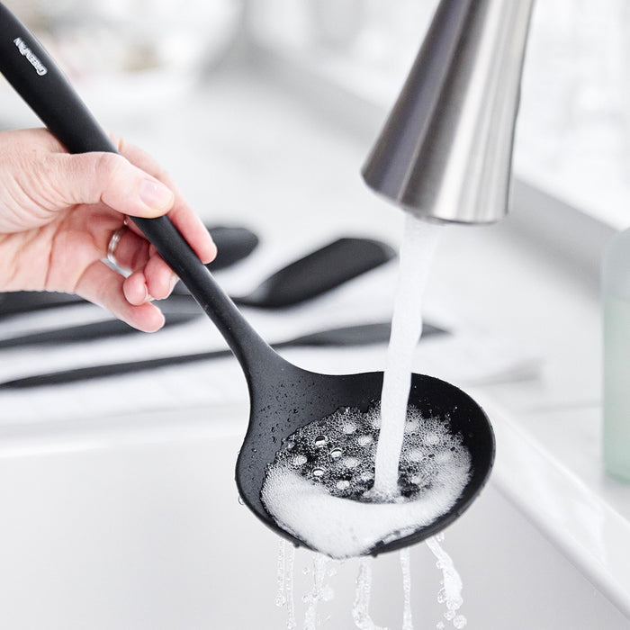Black silicone ladle from 5-piece utensil set being rinsed under running water in a sink with soap bubbles.