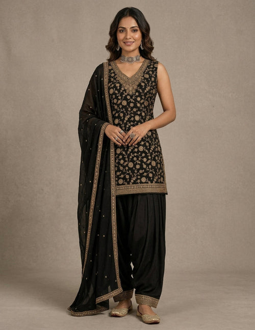 A woman models a traditional black Indian ethnic outfit with intricate gold embroidery, featuring a sleeveless embroidered kurta, black patola-style pants, and a matching black dupatta draped over her shoulder, set against a neutral beige studio background.
