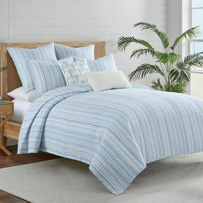 Boracay Stripe Crewel Palm Tree Pillow on a bed with light blue striped bedding and tropical decor.