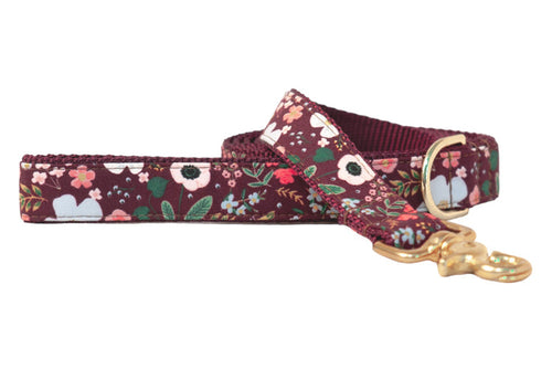 Dog leash with a rich burgundy red fabric  with a variety of floral design of blossoms with leaves. The fabric is sewn onto sturdy burgundy nylon webbing with matching stitching, and the leash is finished with a polished swivel clasp and a matching D-ring near the handle.