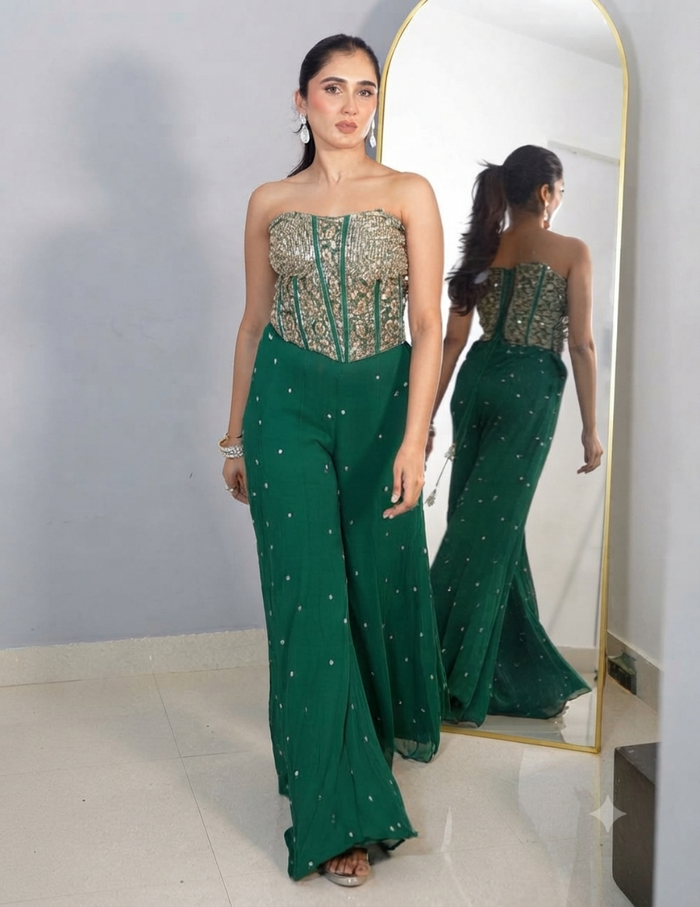 A woman with her hair tied in a ponytail wearing a strapless green embroidered top and matching wide-leg trousers, accessorized with earrings and bracelets, standing in front of a mirror.