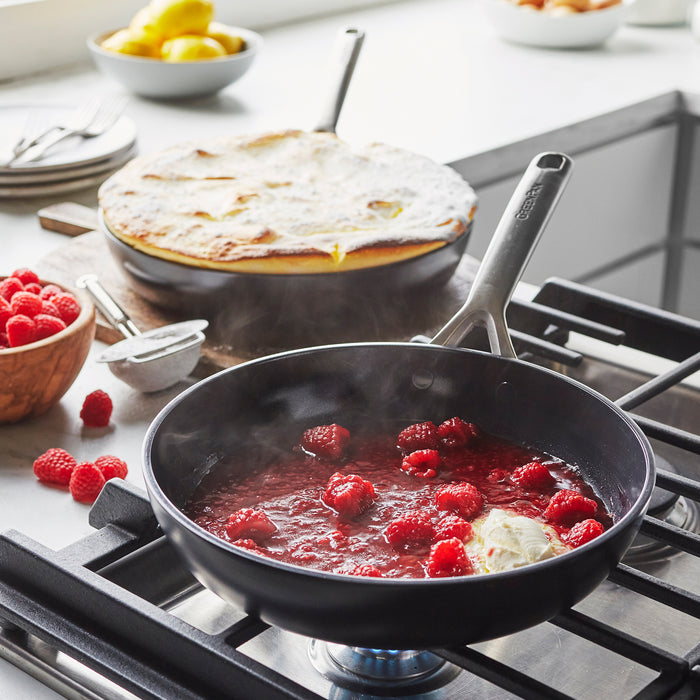 Ceramic nonstick frypan set with crepes cooking, raspberries and sauce on the counter nearby.