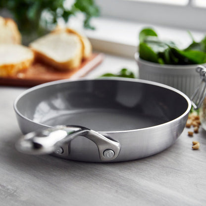 Stainless steel 8-inch frypan with a mirror handle on a kitchen counter, bread and spinach in the background.