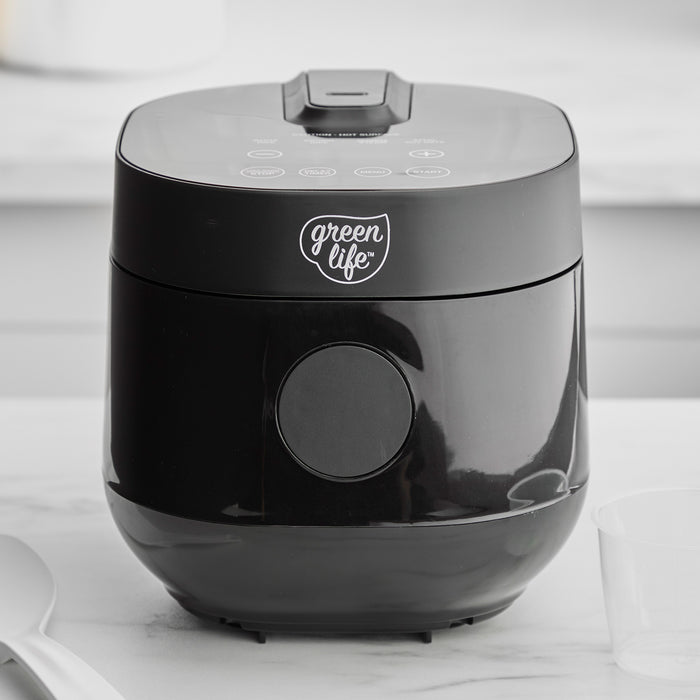 Black 4-cup rice and grains cooker on a white countertop with a spatula and clear measuring cup beside it.