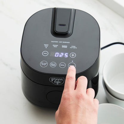 Hand pressing button on black GreenLife 4-cup rice and grain cooker, digital timer display shows 25 minutes.