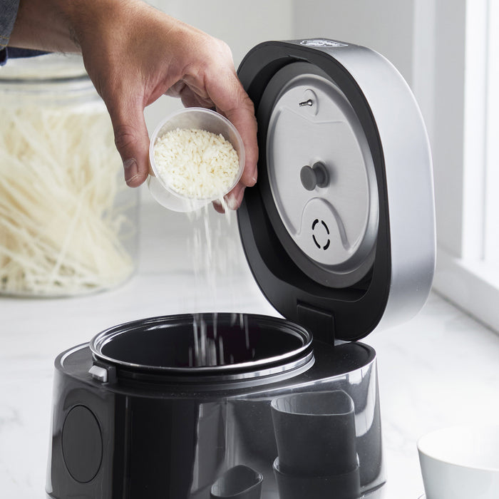 White rice being poured into an open black GreenLife 4-cup rice and grains cooker on a kitchen counter.