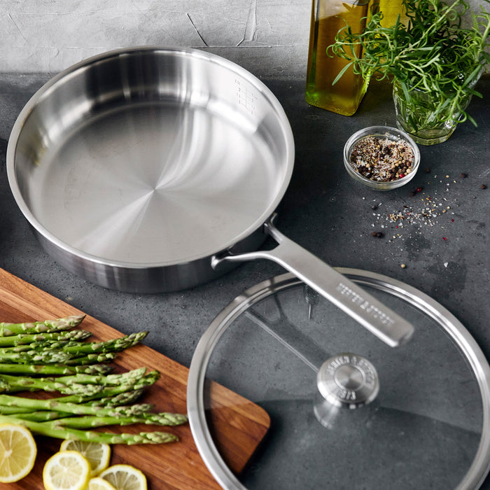 Stainless steel sauté pan with lid on a counter, shown with asparagus spears and lemon slices nearby.