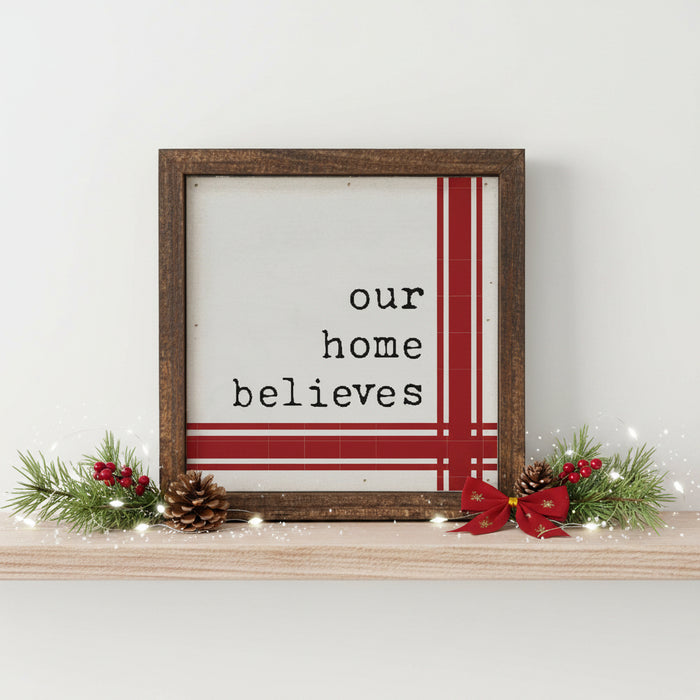 Our Home Believes Vintage Christmas Sign 6x6 A square framed wall sign with a rustic dark brown wooden frame. The sign features a white background with the typewriter-style text 'our home believes' in black, accented by red vertical and horizontal stripes on the right and bottom edges. Displayed on a wood shelf with pine boughts and red ribbon.