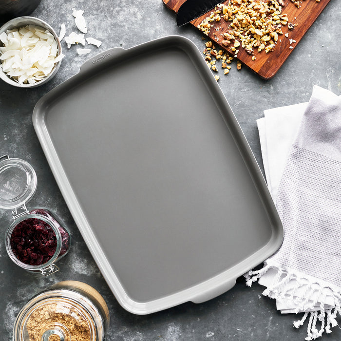 Gray ceramic nonstick half sheet pan with nuts, cranberries, coconut, and oats on a marble surface.