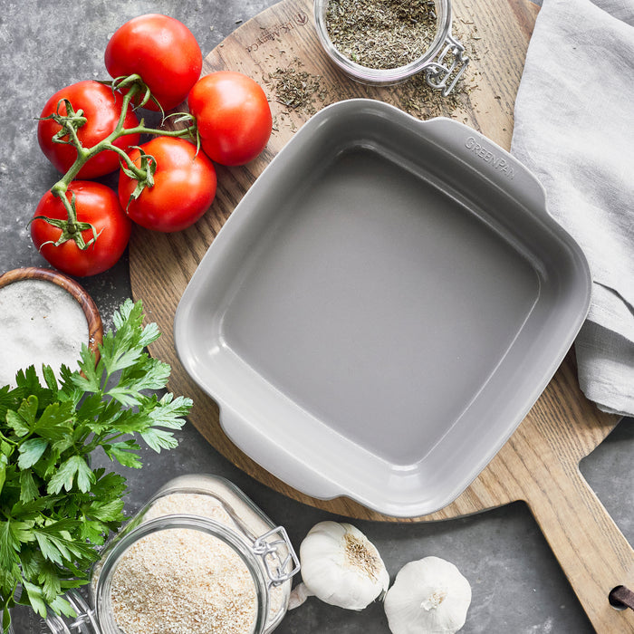 Gray ceramic nonstick square baker on a wooden board, surrounded by tomatoes, parsley, garlic, and herbs.