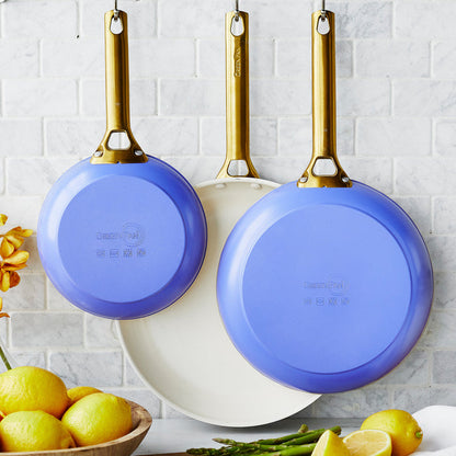 Set of wisteria-colored ceramic nonstick frypans hanging on a tiled wall above lemons and asparagus.