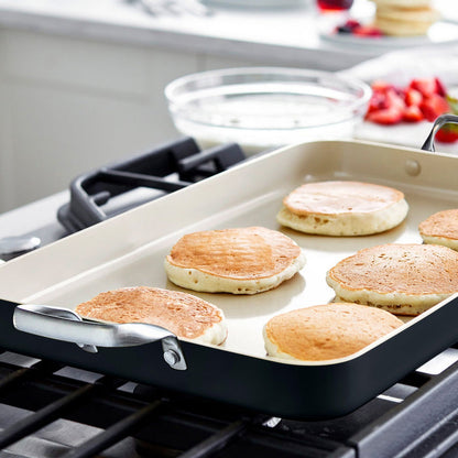 Six pancakes cooking on a black double griddle with bowls and ingredients in the background.