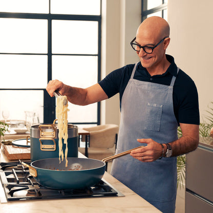 Man in glasses and apron lifts pasta using Stanley Tucci ceramic nonstick cookware set with multiple pieces.