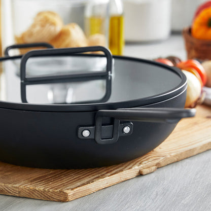 Black ceramic 6.5-quart pan with handles on a wooden board, assorted vegetables in the background.