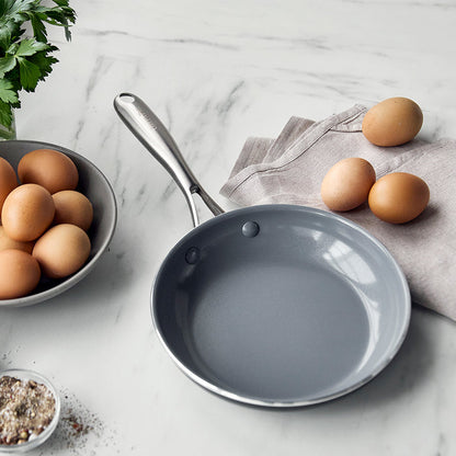 Ceramic nonstick 8-inch frypan next to brown eggs, fresh herbs, gray napkin, and a small dish of seasoning.
