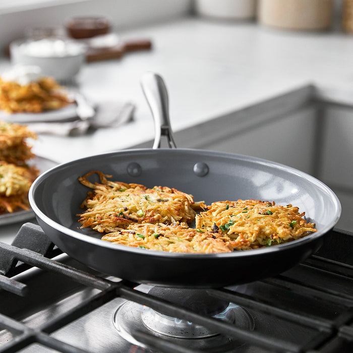 Three potato pancakes cooking in a Swift ceramic nonstick frypan, with more pancakes stacked on a plate in the background.