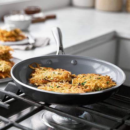 Three potato pancakes cooking in a Swift ceramic nonstick frypan, with more pancakes stacked on a plate in the background.