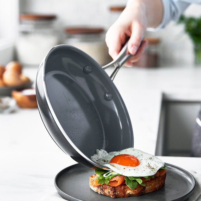 A hand slides a fried egg from a gray ceramic nonstick frypan onto toast with greens on a gray plate.