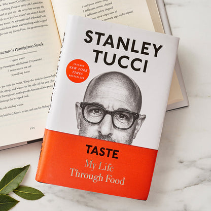 Stanley Tucci’s Taste: My Life Through Food cookbook open on a white surface with green leaves nearby.