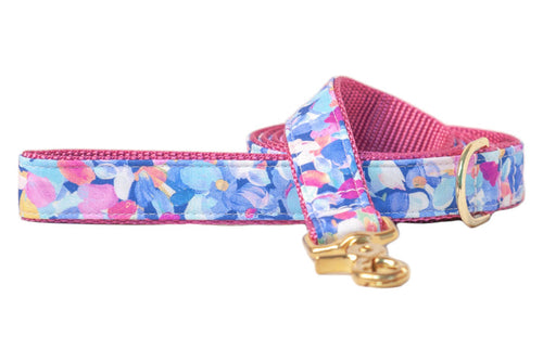 A stylish dog leash featuring a vivid floral print with overlapping petals in shades of blue, pink, purple, yellow, and white. The outer layer is smooth and patterned, while the inner side is made of textured pink woven nylon for durability and comfort. It includes a sturdy gold-colored metal swivel snap hook and a matching D-ring for added attachment options.